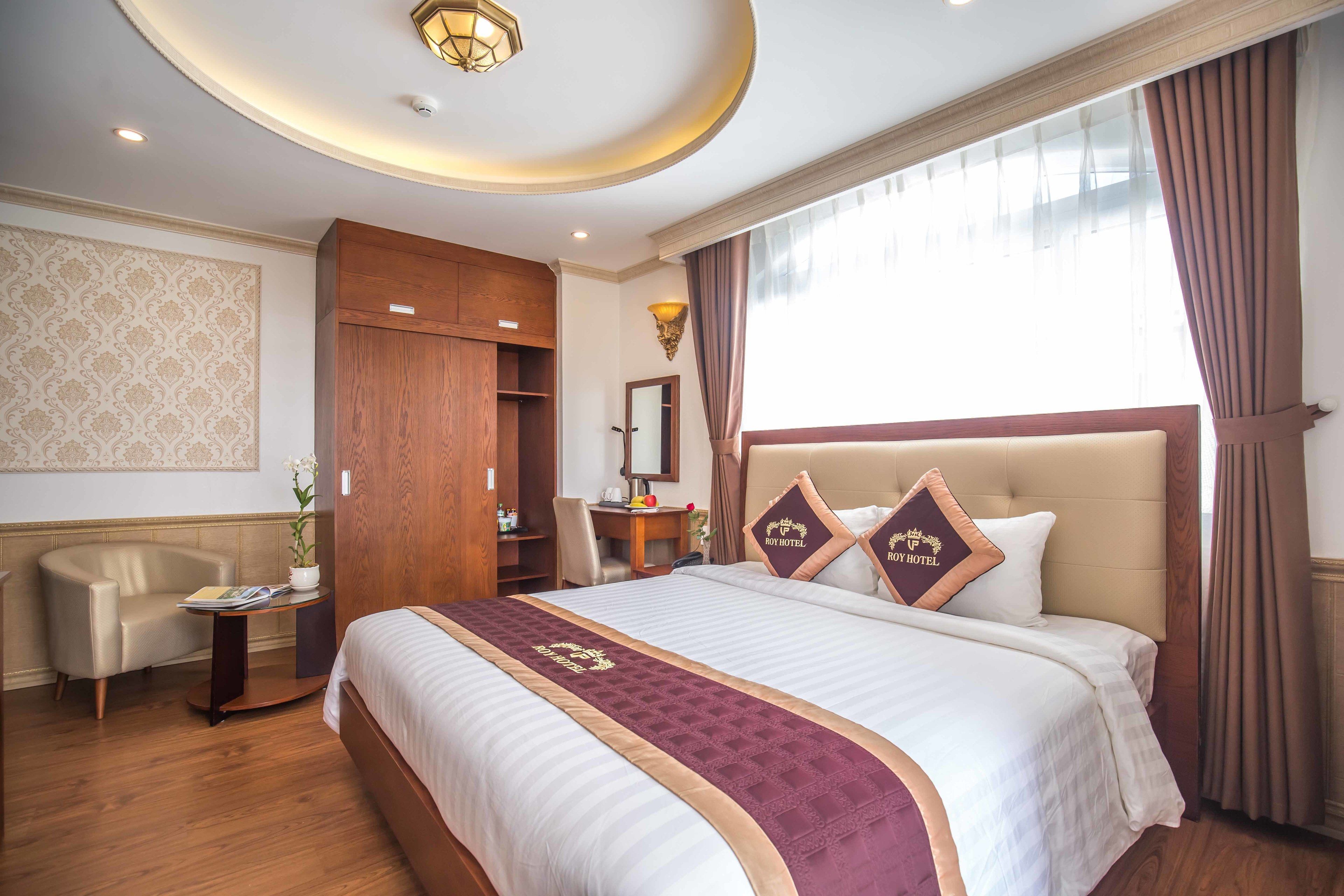 Une chambre d’hôtel avec un grand lit, une armoire en bois, un bureau avec une chaise, une petite table avec une plante et une grande fenêtre avec des rideaux.