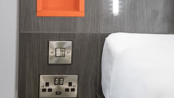 A modern hotel room with a grey wood panel headboard, an orange shelf, a bedside switch, and a power outlet.