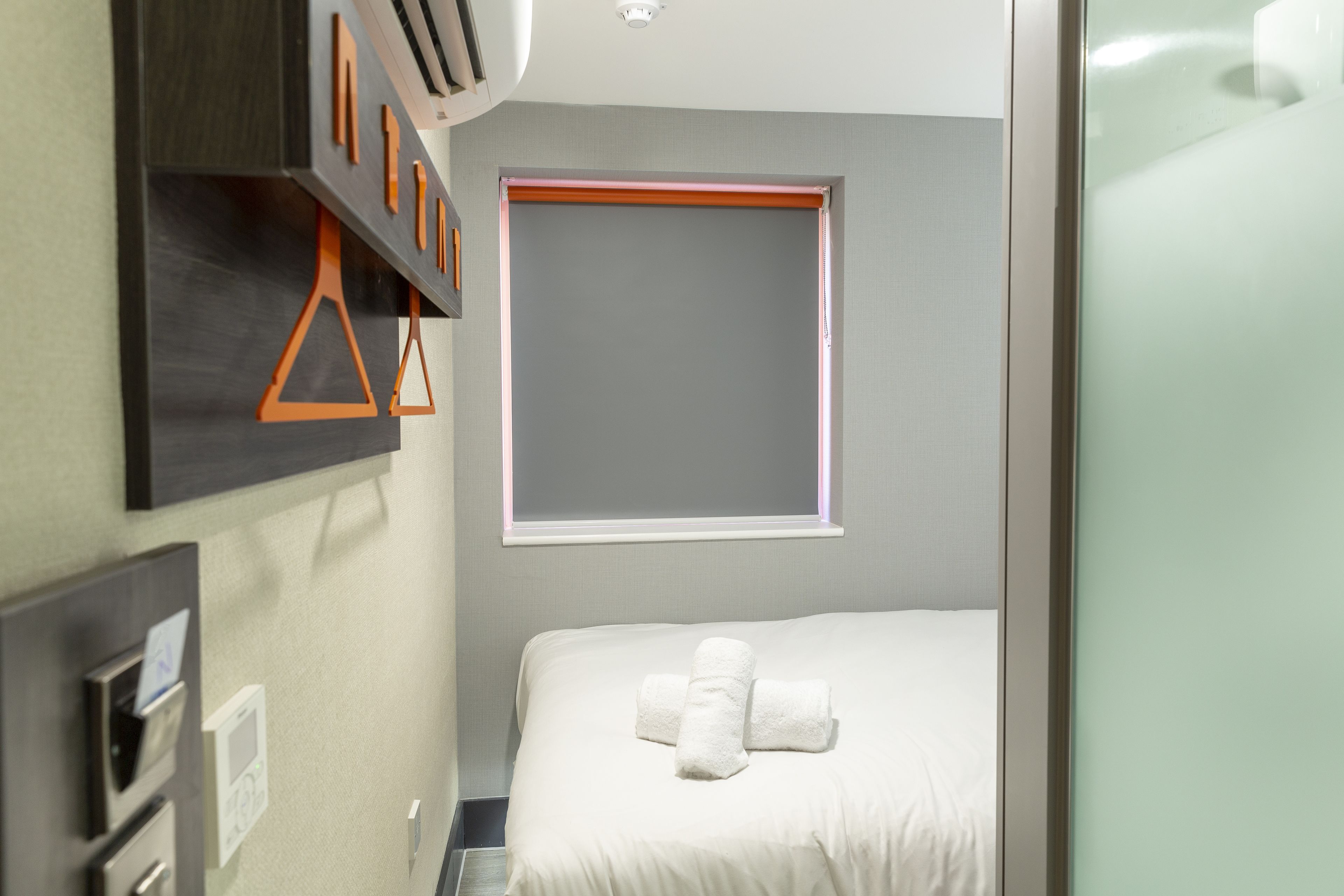 A hotel room with a bed, a window, a wall-mounted air conditioning unit, and a towel rack.