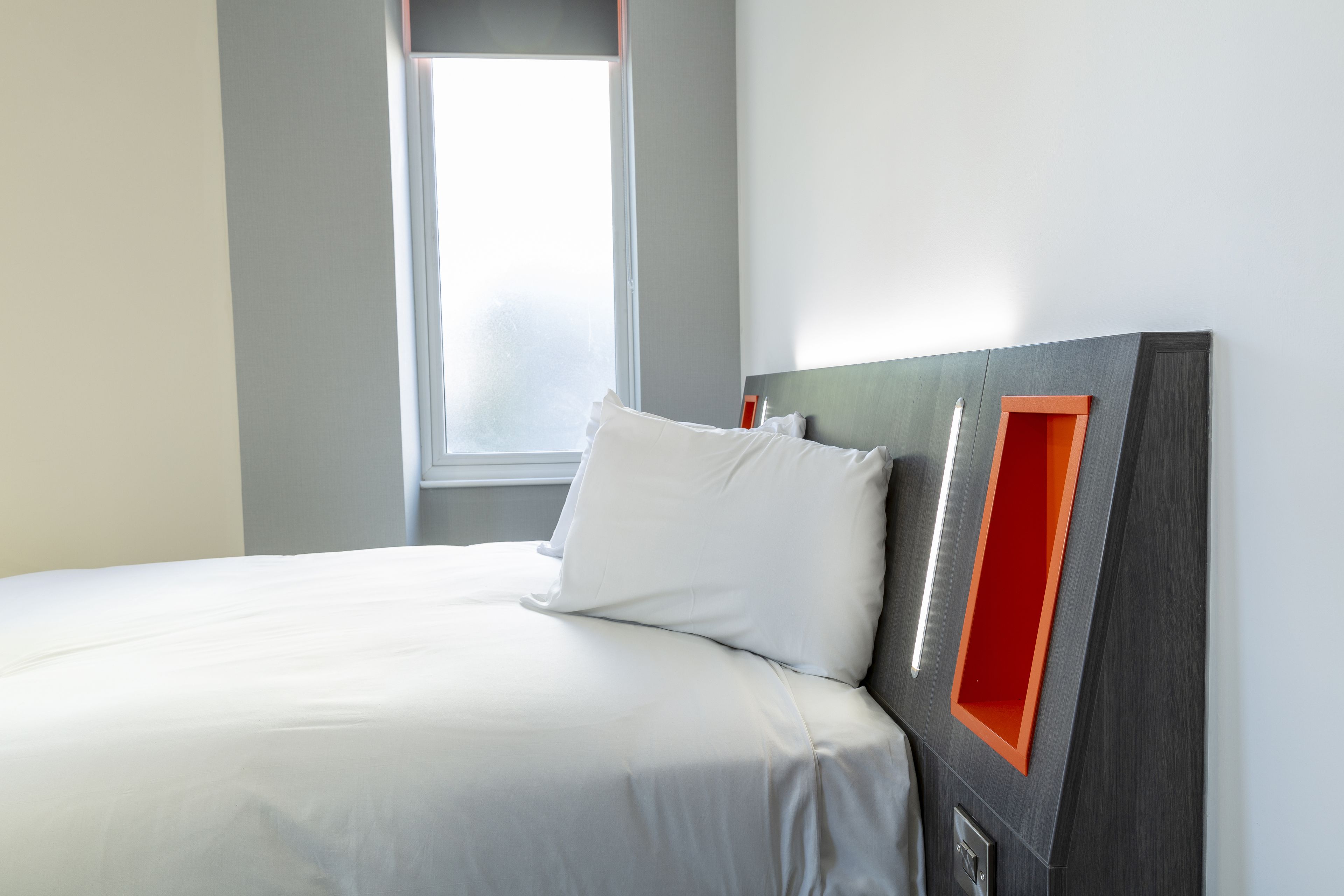 A neatly made bed with white linens, a modern headboard with a red accent, and a window providing natural light.