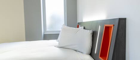 A neatly made bed with white linens, a modern headboard with a red accent, and a window providing natural light.