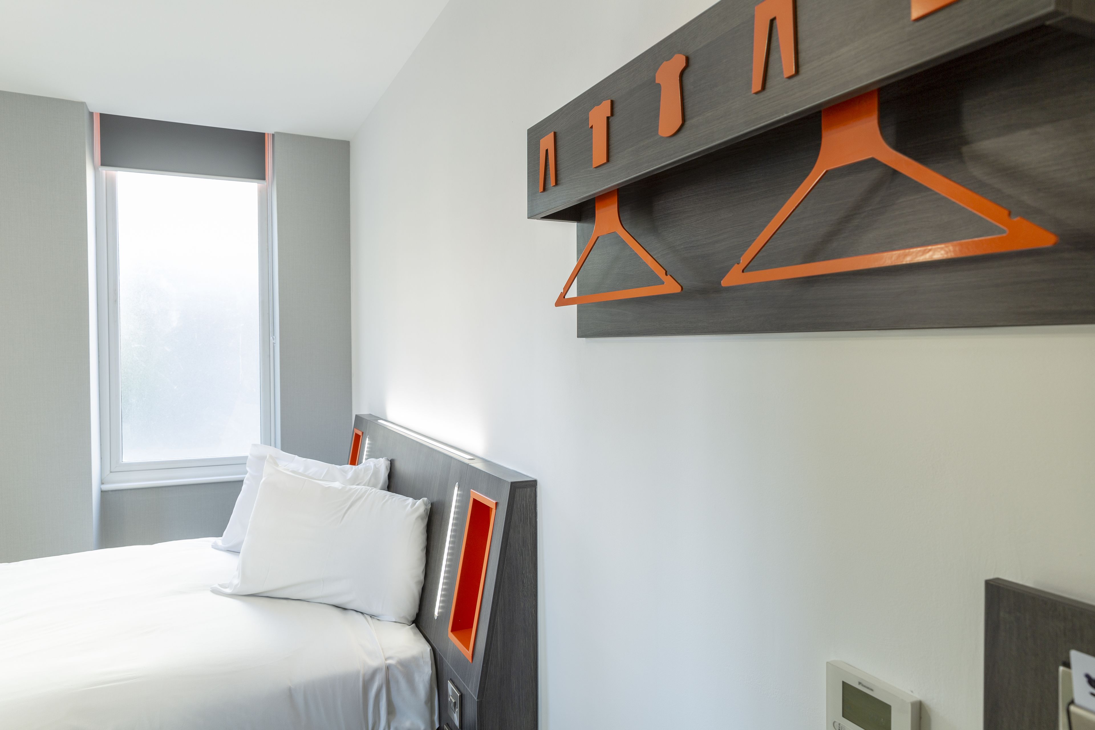 A modern bedroom with a bed, a window, and a wall-mounted storage unit with orange hangers.