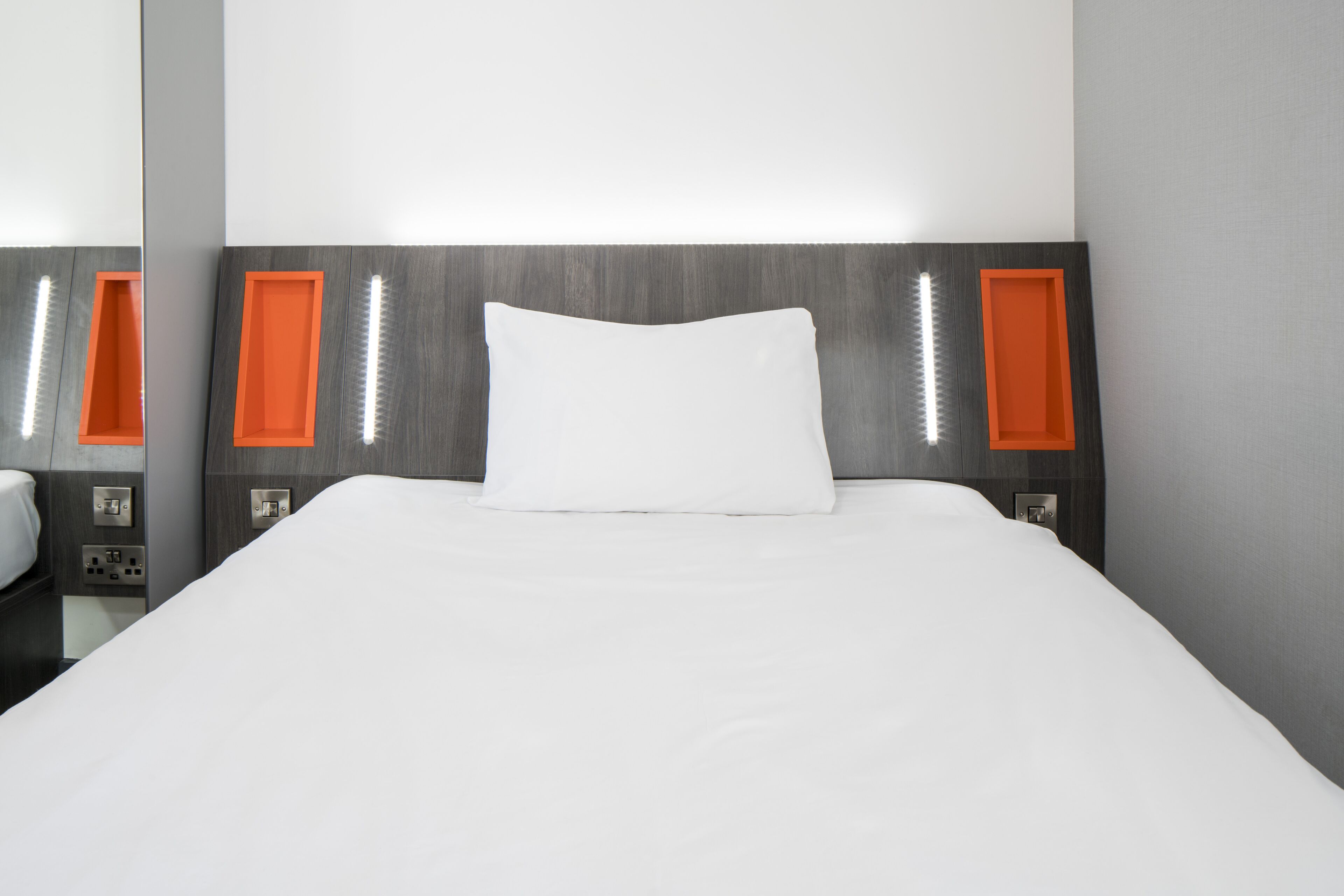 A neatly made bed with a white pillow and a grey headboard with orange accents.