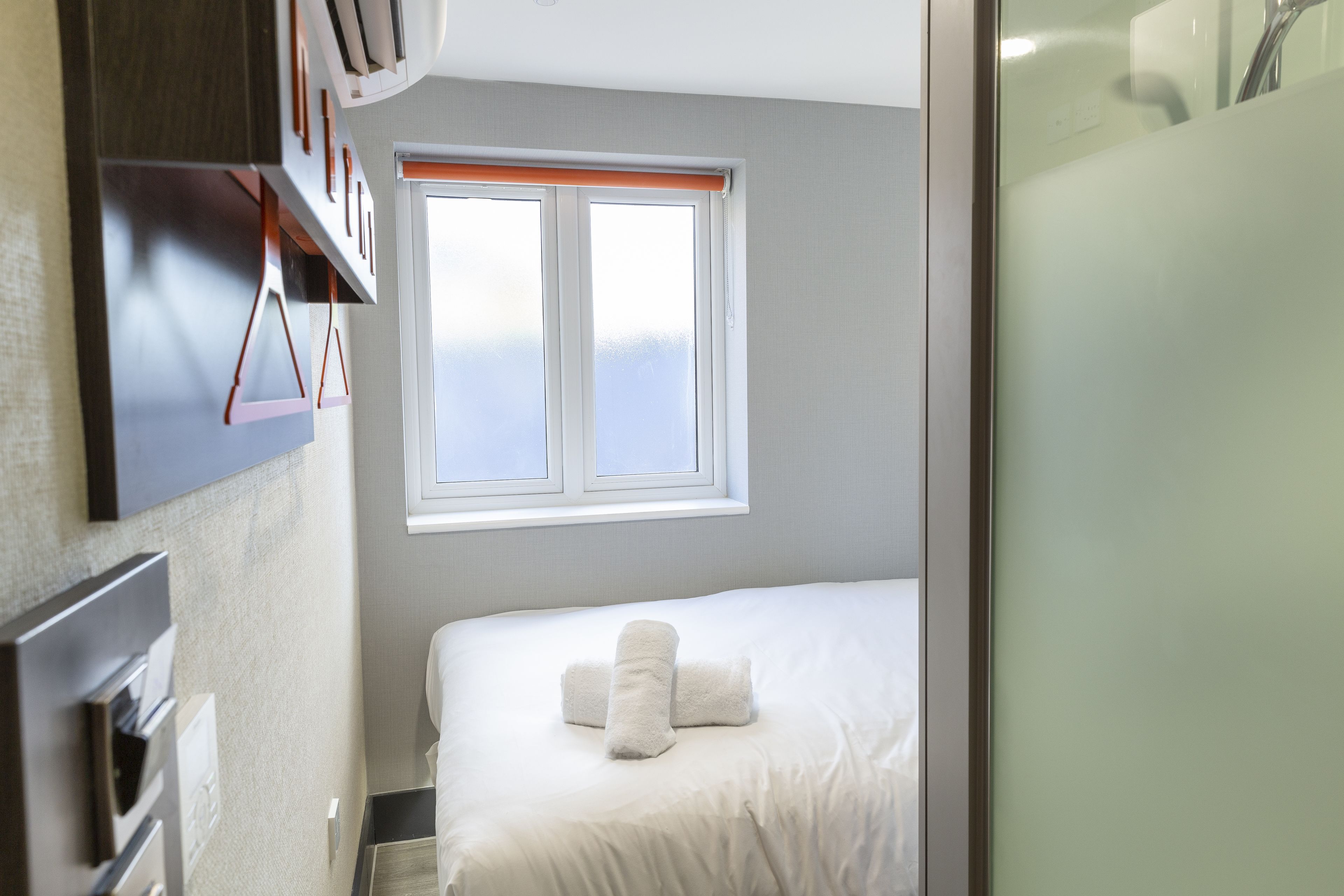 Hotellihuone, jossa on sänky, ikkuna ja seinälle kiinnitetty hylly.