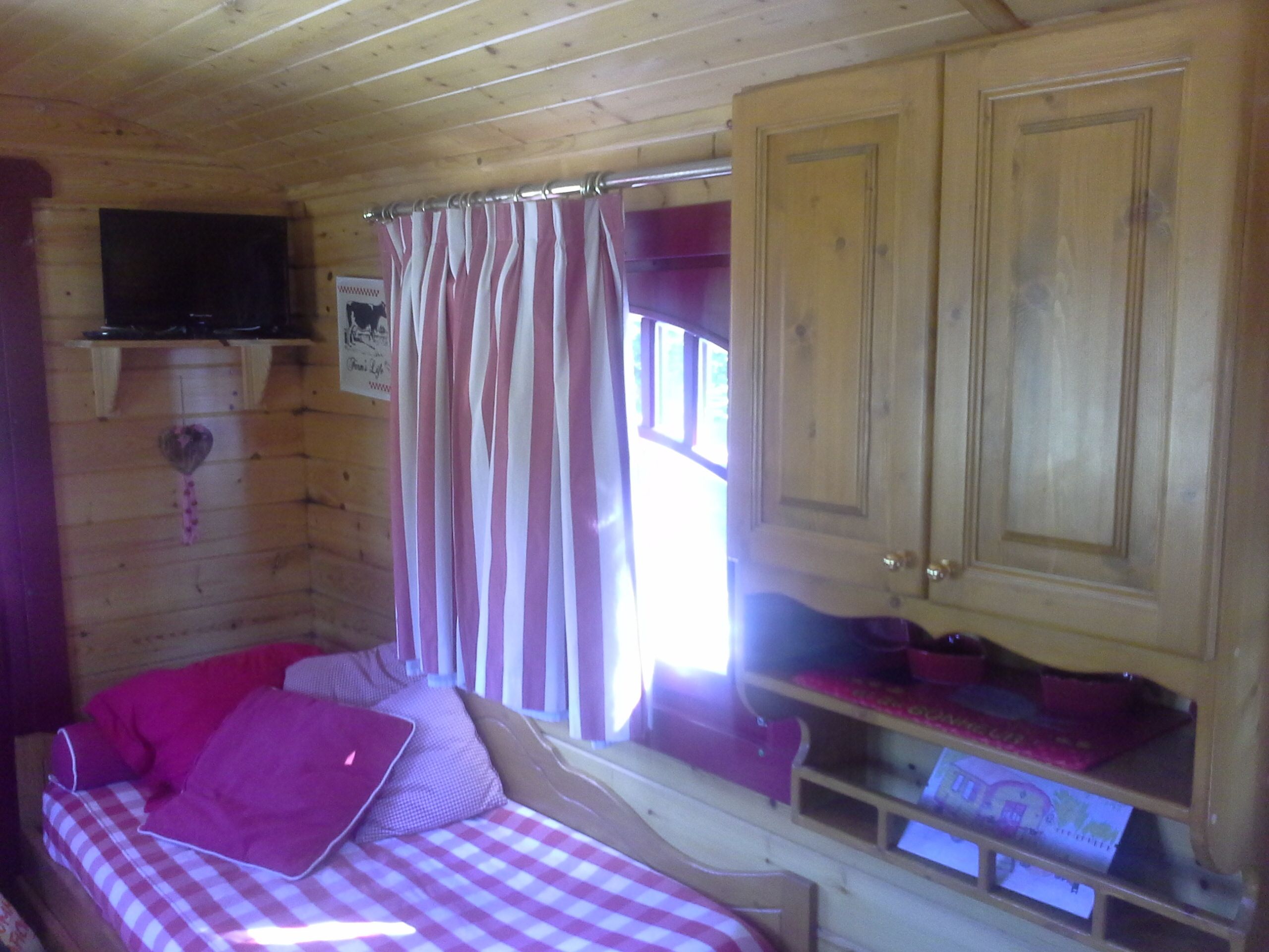 A wooden cabin room with a bed, a television mounted on the wall, and a window with curtains.