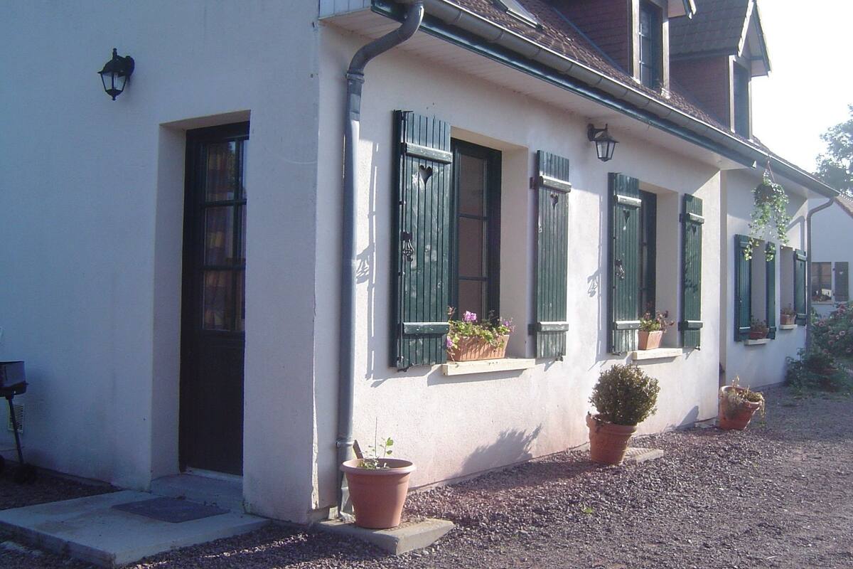 A white building with green shutters, potted plants, and a gravel driveway.