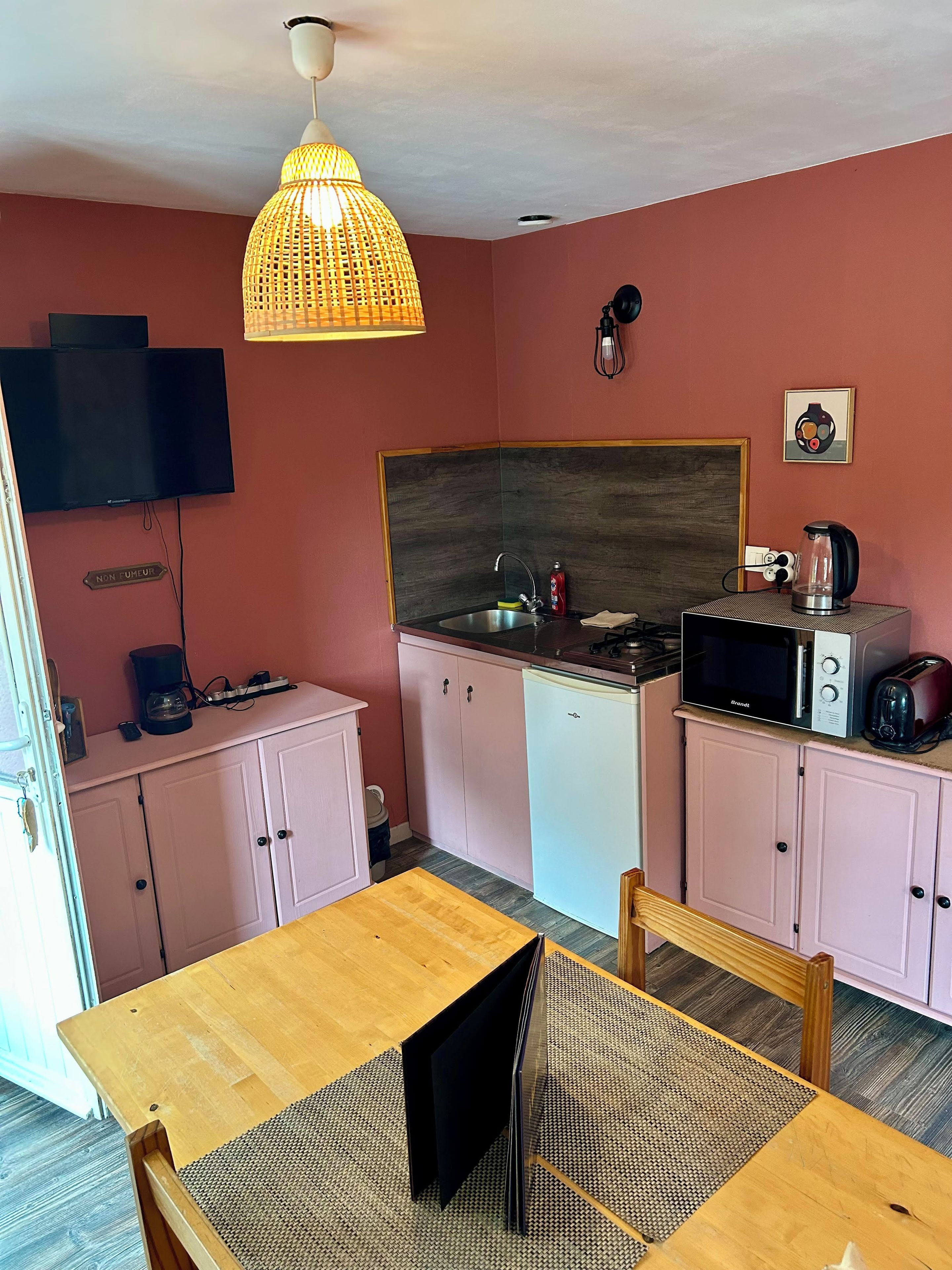 A compact kitchen with a microwave, toaster, and coffee maker, a television mounted on the wall, and a wooden dining table with chairs.