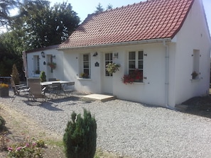A white building with a red-tiled roof, a gravel driveway, and a small patio with a table and chairs. - Roulotte et Gîte chambres d'hôtes (Saint-Pol-sur-Ternoise)