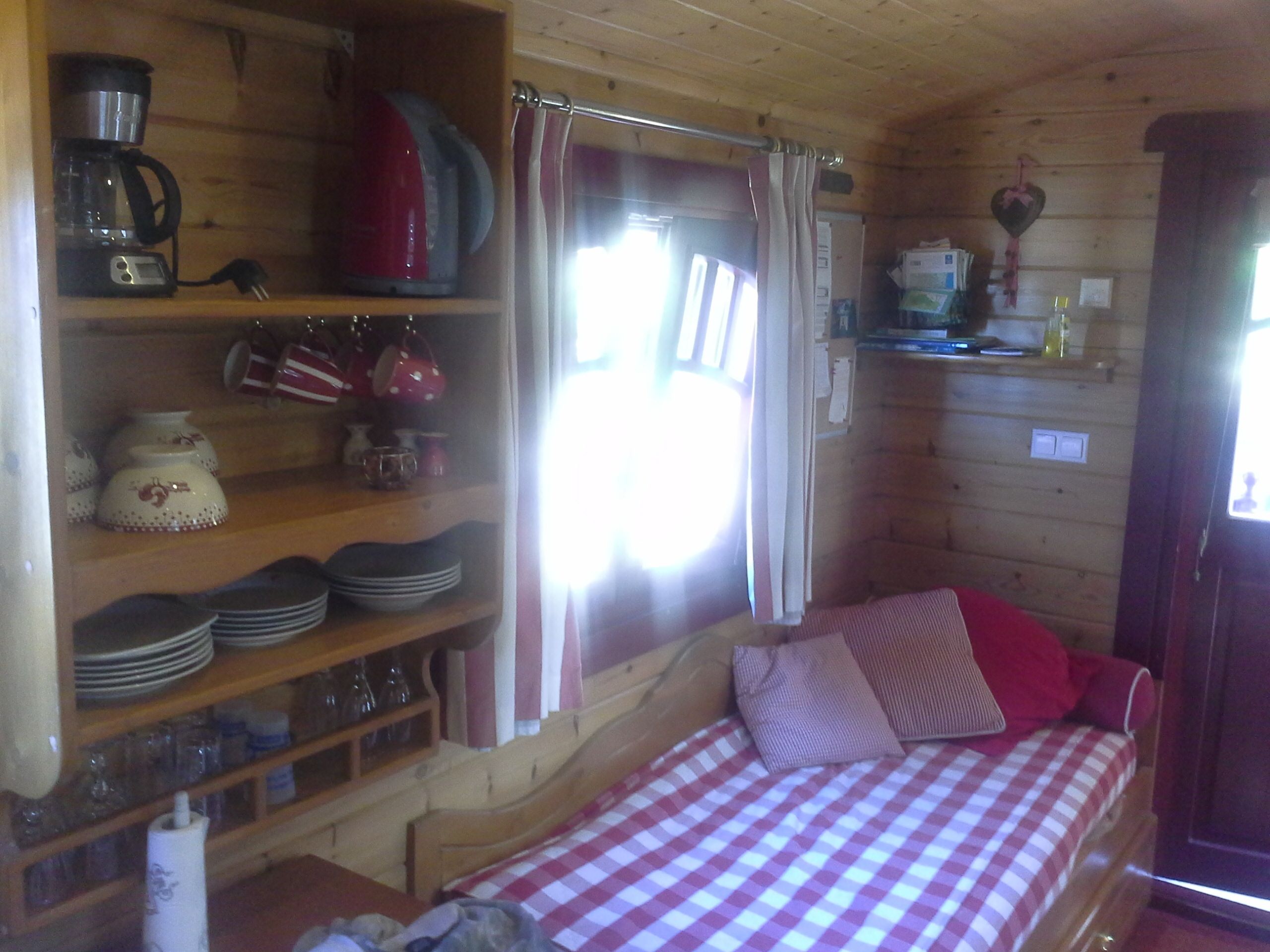 Interior acogedor de una cabaña con una cama, estantes con utensilios de cocina, una cafetera y una ventana con cortinas.