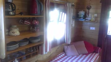 Interior acogedor de una cabaña con una cama, estantes con utensilios de cocina, una cafetera y una ventana con cortinas.