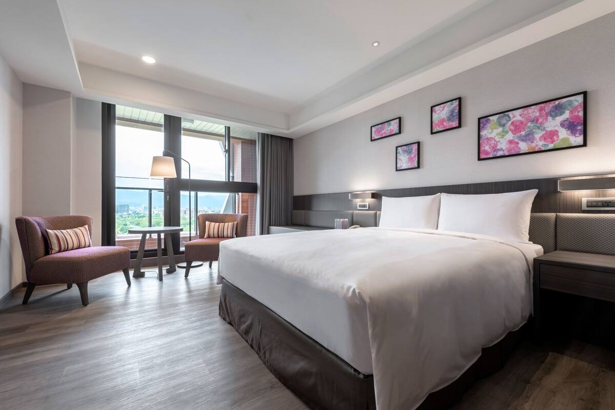 Ett modernt hotellrum med en stor säng, en sittgrupp med två fåtöljer, ett litet bord och ett stort fönster med utsikt över staden.