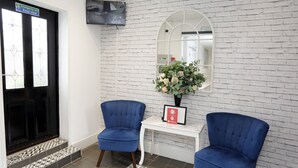A reception area with a white brick wall, a black door with a glass panel, two blue chairs, a small white table with a vase of flowers, and a mounted television. - Bexley Village Hotel (Bexley)