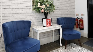 A reception area with white brick walls, two blue chairs, a small white table with a vase of flowers, a framed certificate, and a fire extinguisher. - Bexley Village Hotel (Bexley)