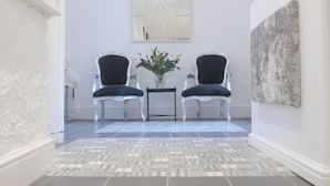 A hallway with patterned floor tiles, two black upholstered chairs, a small table with a vase of flowers, and a large abstract painting on the wall. - Bexley Village Hotel (Bexley)