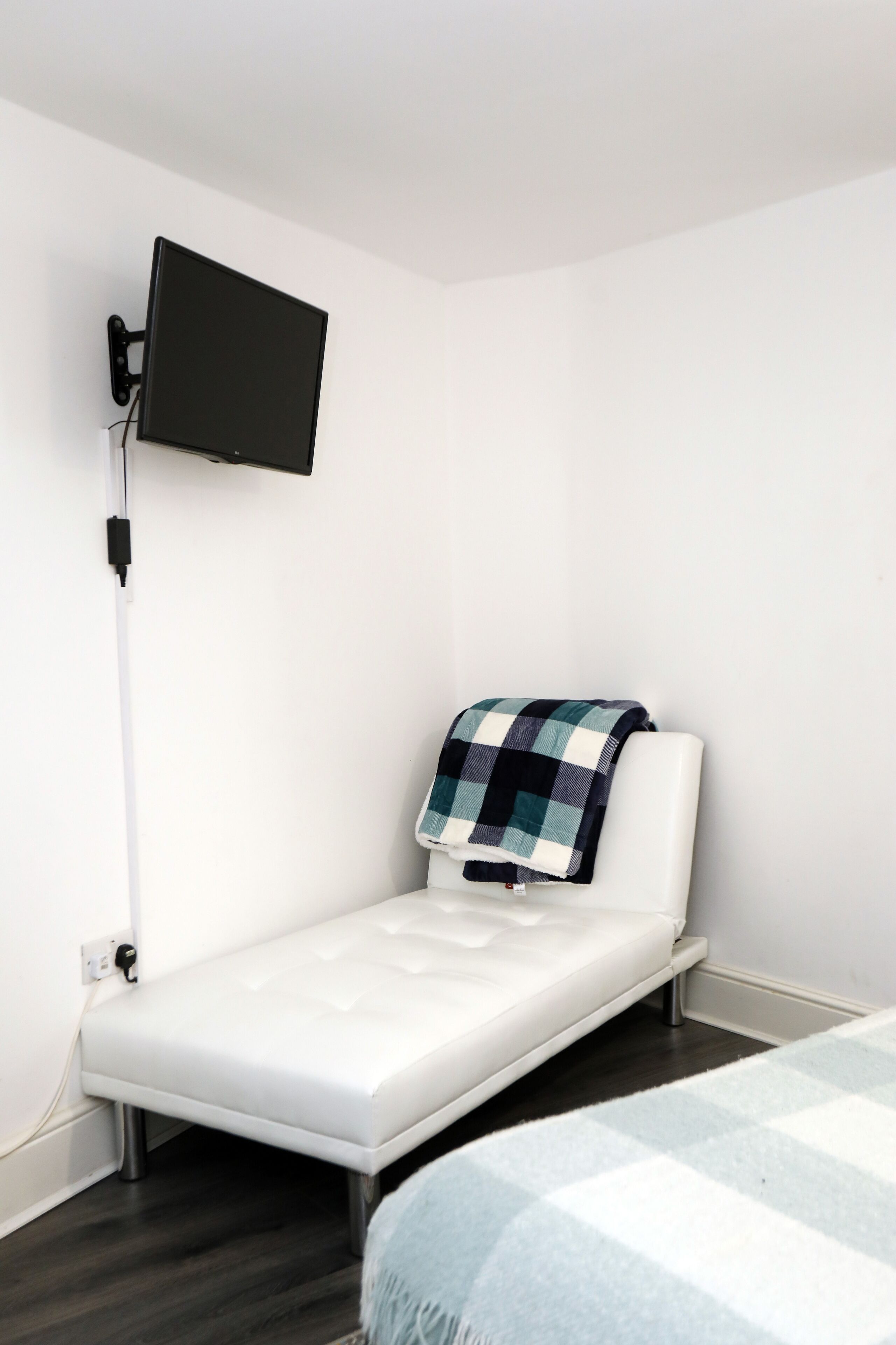 Ein kleines Zimmer mit einem Einzelbett, einem an der Wand montierten Fernseher, einem Nachttisch und einer Decke auf dem Bett.