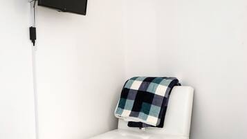 Ein kleines Zimmer mit einem Einzelbett, einem an der Wand montierten Fernseher, einem Nachttisch und einer Decke auf dem Bett.