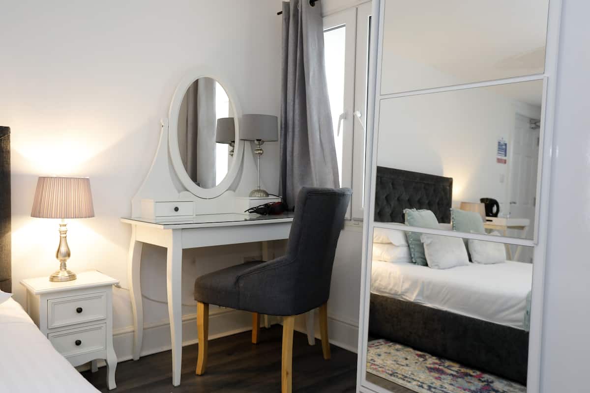A bedroom with a bed, a desk with a mirror, a chair, a bedside table, and a nightstand.