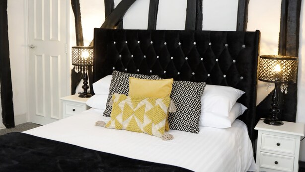 A bedroom with a black and white Tudor-style headboard, a white bed with multiple pillows, two bedside tables with lamps, and a white door. - Bexley Village Hotel (Bexley)