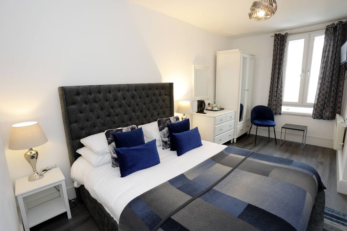 A neatly arranged bedroom with a large bed, a bedside table with a lamp, a wardrobe, a chair, and a window with curtains.