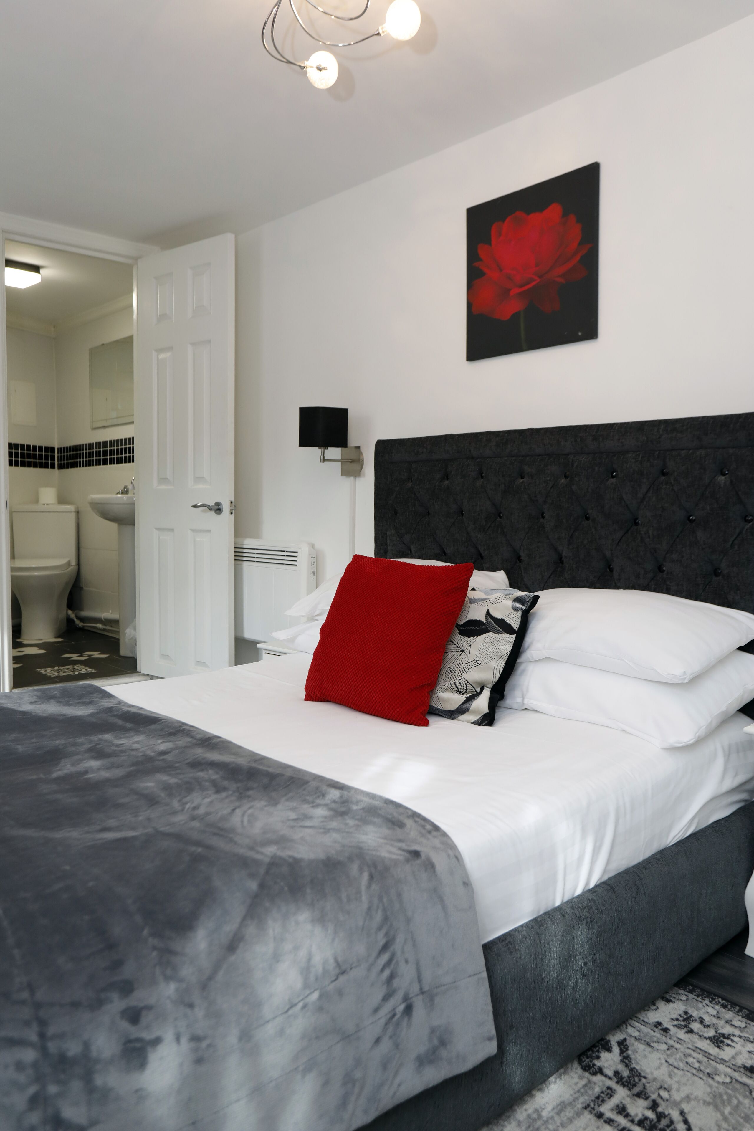 Un dormitorio con cama, almohada roja, almohada blanco y negro, luz empotrada en la pared y un cuadro con una flor roja.