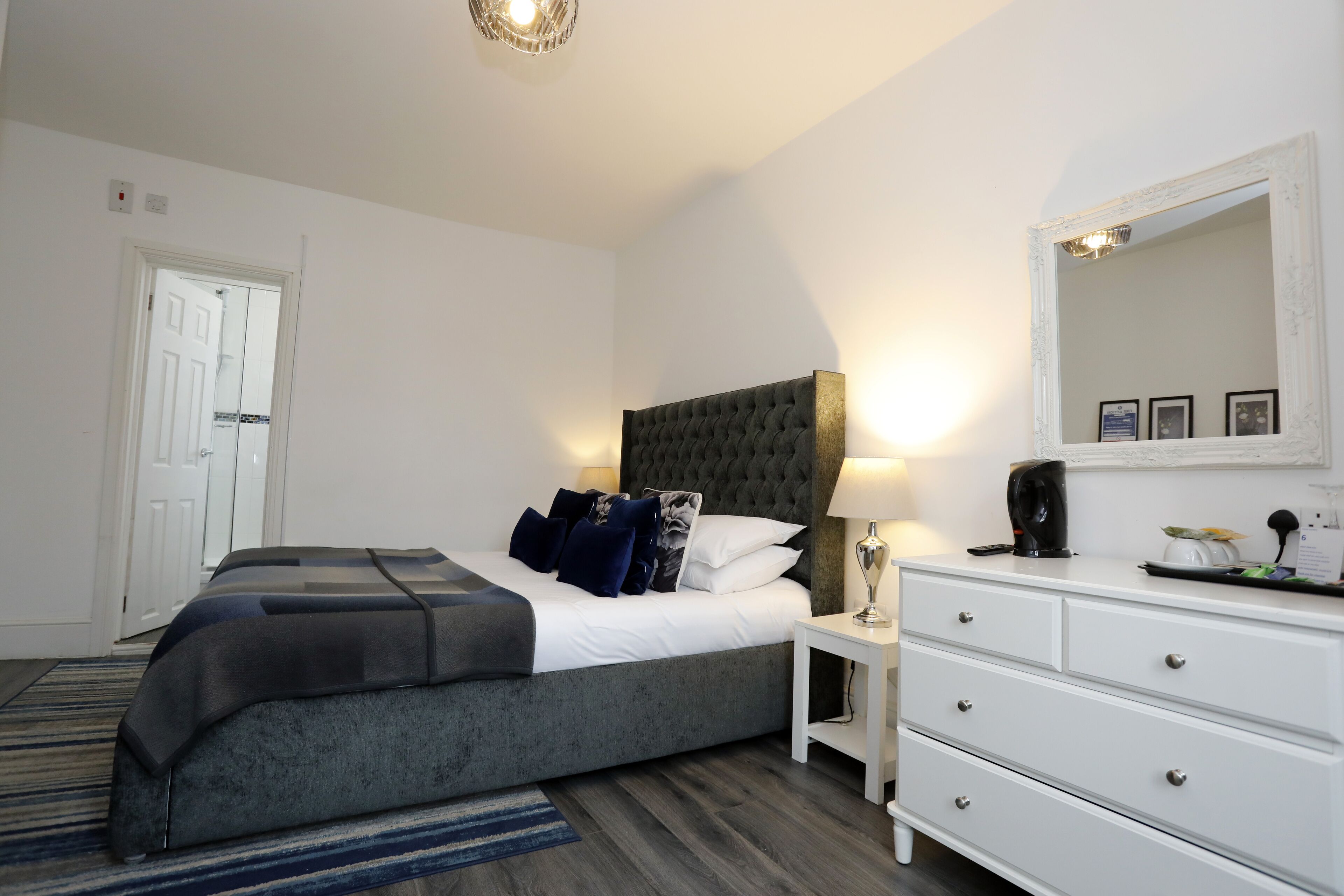 A bedroom with a large bed, a bedside table with a lamp, a dresser, and a mirror on the wall.