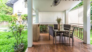 Eine überdachte Terrasse mit Tisch und Stühlen, umgeben von Grünpflanzen und Blick auf einen Garten.