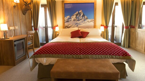 A hotel room with a large bed, a wooden headboard, a mounted deer head, a mountain painting, and a desk with a lamp. - Marmore (Valtournenche)
