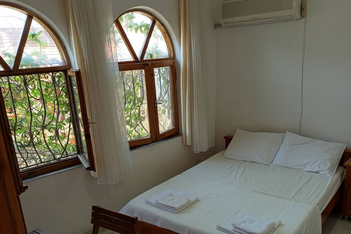 A bedroom with a bed, two windows with curtains, a table with a chair, and an air conditioning unit.