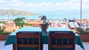 Un balcon avec une table et des chaises, donnant sur une marina où sont amarrés des bateaux, et sur un paysage vallonné.