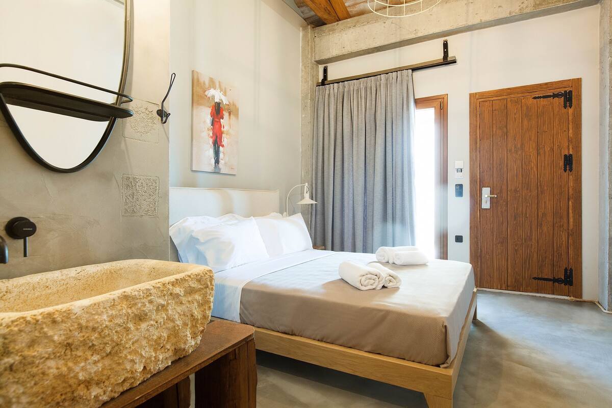 A bedroom with a bed, a wooden bedside table, a stone sink, a painting, a window with curtains, and a wooden door.