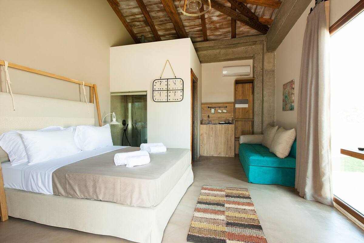 A bedroom with a bed, a wooden headboard, a hanging light fixture, a kitchen area with wooden cabinets, and a teal sofa.