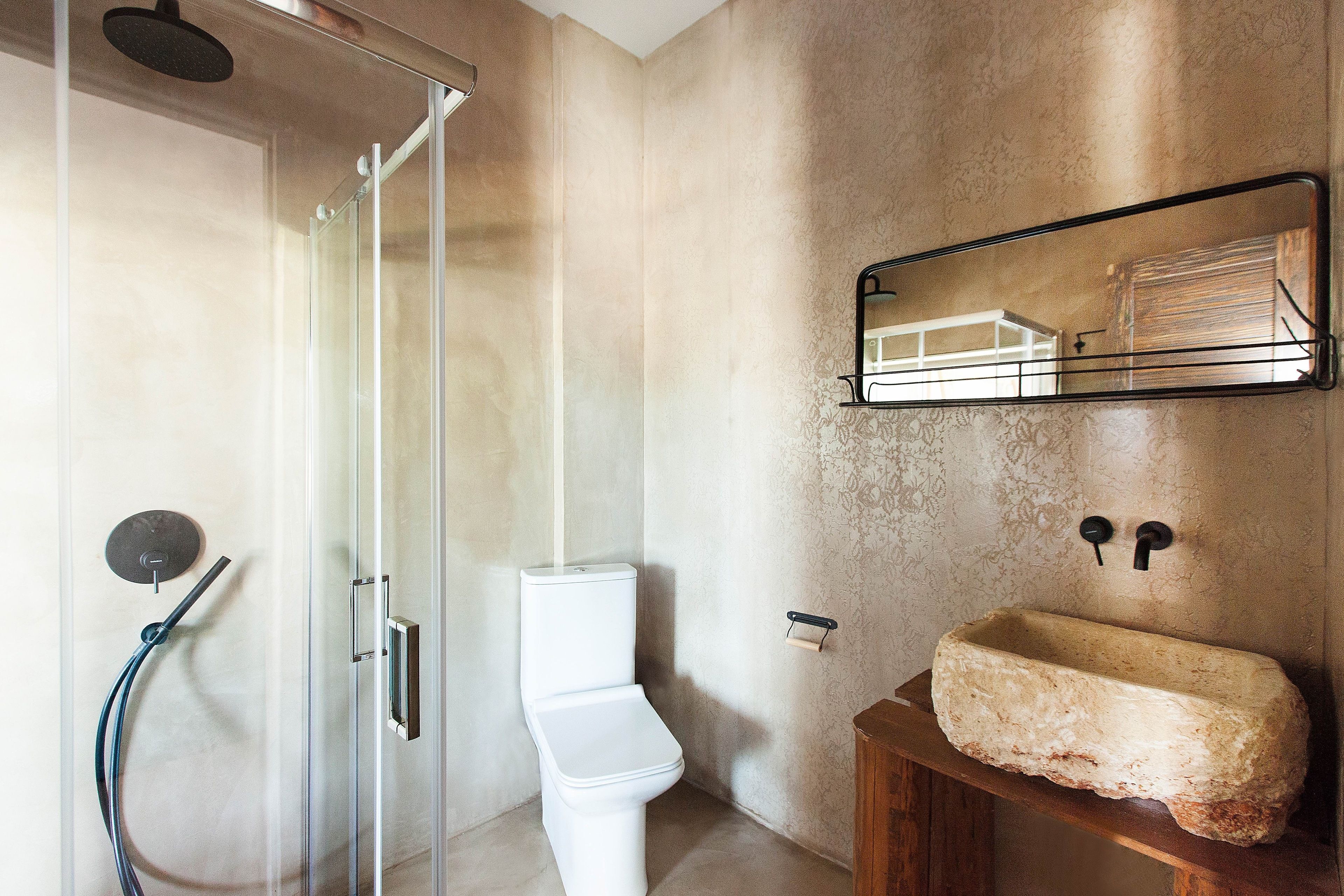 A bathroom with a glass-enclosed shower, a white toilet, and a stone sink on a wooden shelf.