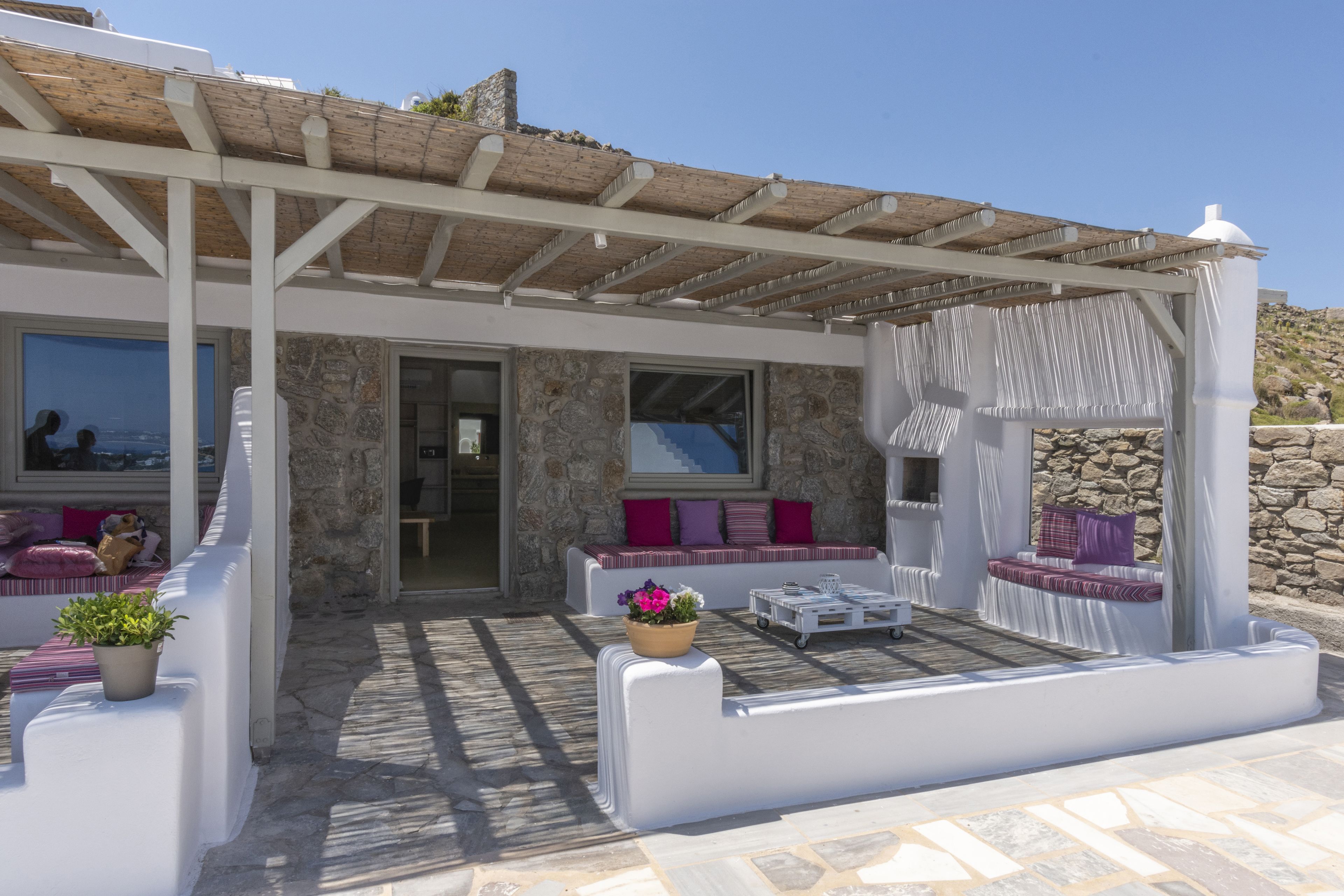 Un espacio de estar al aire libre cubierto, con paredes de piedra, muebles blancos y vista al mar.