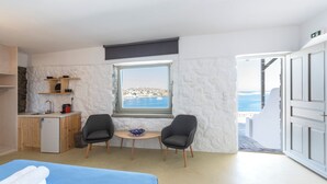 A room with a bed, a small kitchenette, a window with a view of the sea, and two chairs. - Mykonos Azure (Mykonos)