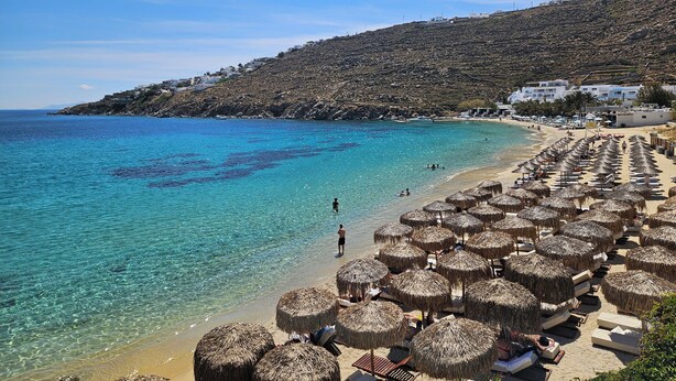 A beach with clear turquoise water, thatched umbrellas, and a hillside with buildings. - Mykonos Azure (Mykonos)
