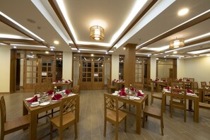 A dining area with multiple tables set for service, wooden chairs, and a modern ceiling design with recessed lighting.