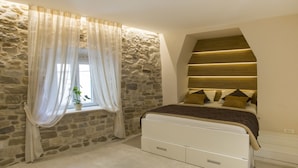 Une chambre avec un mur en pierre, un lit doté d’une tête de lit et une fenêtre ornée de rideaux légers.