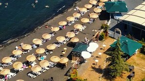 An aerial view of a beach with rows of umbrellas and sun loungers, a rocky breakwater, and a few people swimming in the water. - Mare Blu Hotel (Akçaabat)