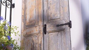 A rustic wooden door with metal hinges and a latch, set against a white wall with a black metal gate to the left. - Blue Jasmine Suites (Rhodes)