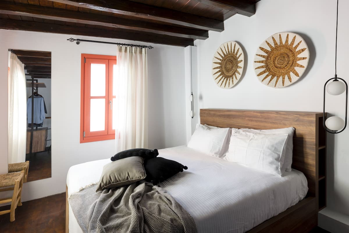Un dormitorio con cabecera de madera, cama con ropa blanca, dos piezas decorativas que evocan el sol en la pared, una ventana con contraventanas naranjas y una silla de madera.