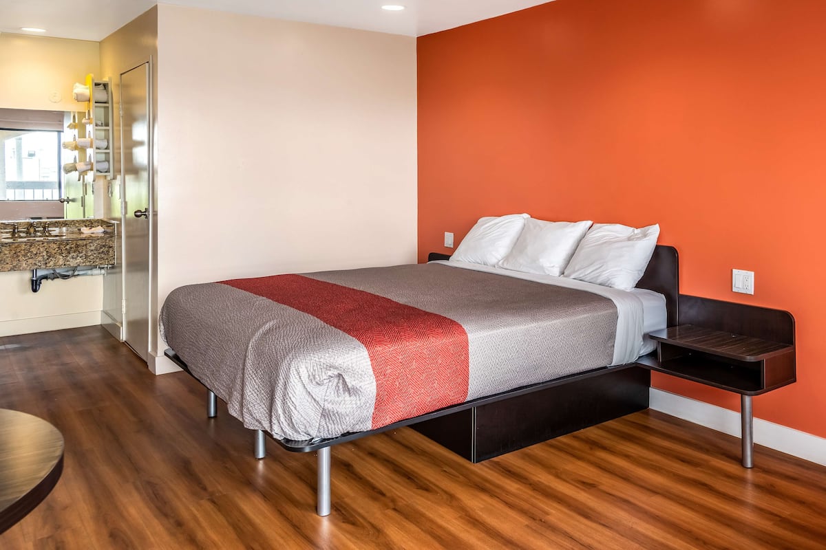 A modern hotel room with a bed, a wooden floor, a bathroom with a mirror, and a red accent wall.