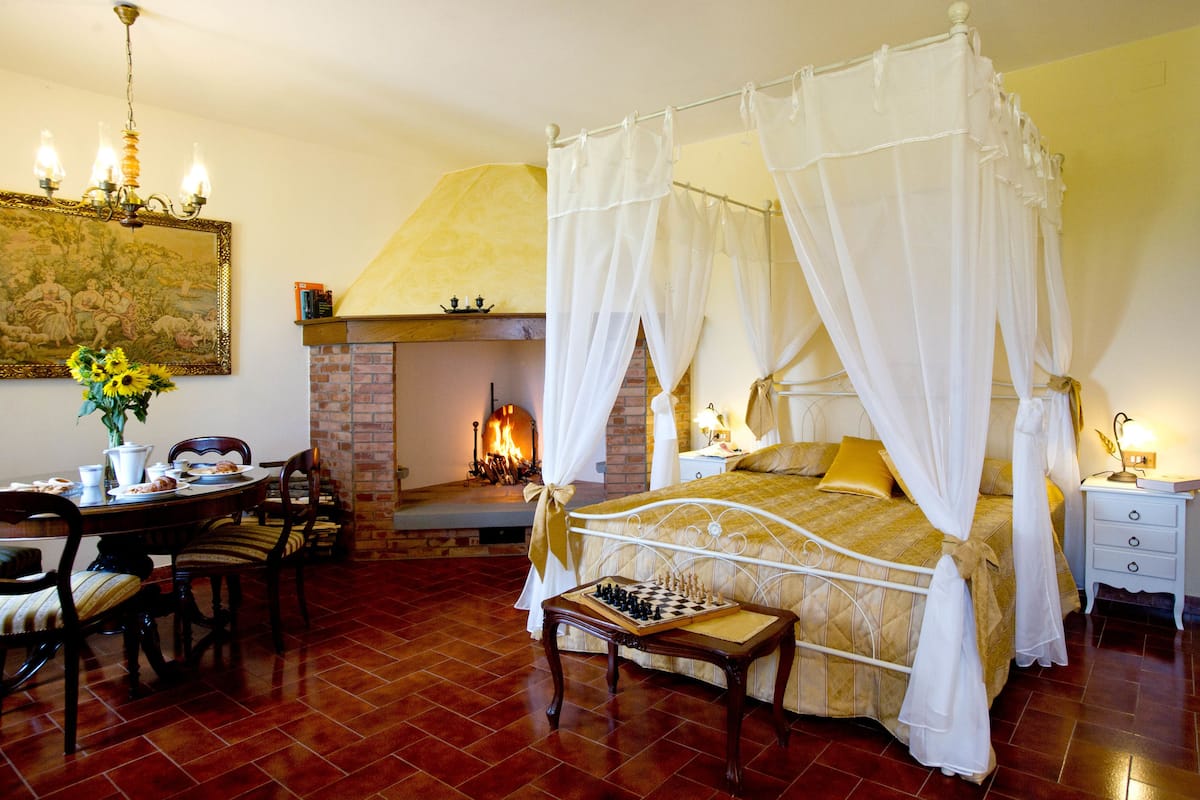 A four-poster bed with a canopy, a fireplace, a chessboard, and a dining area with chairs and a table.