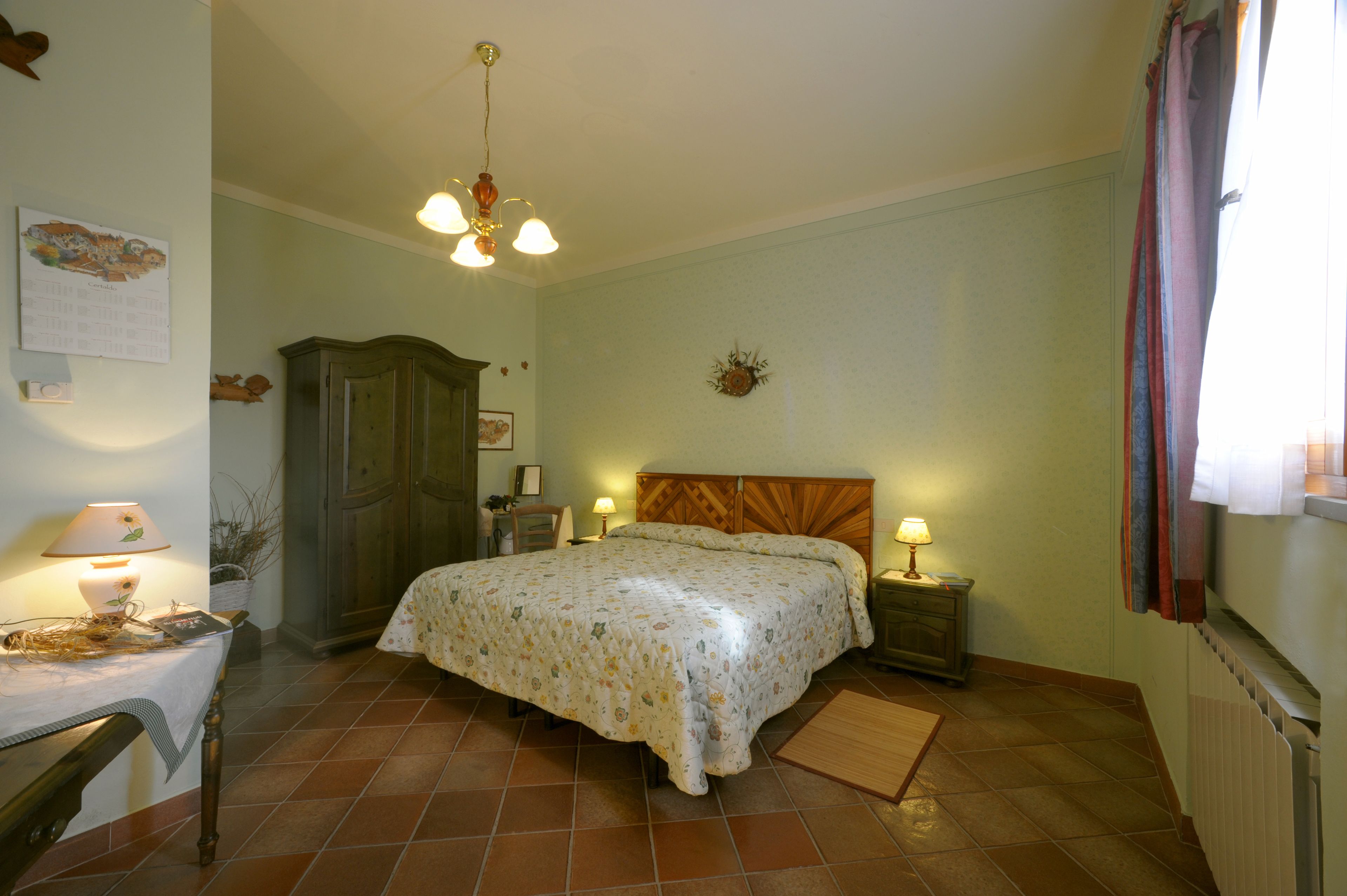 A bedroom with a bed, nightstands, a wooden wardrobe, a desk with a lamp, and a wall calendar.