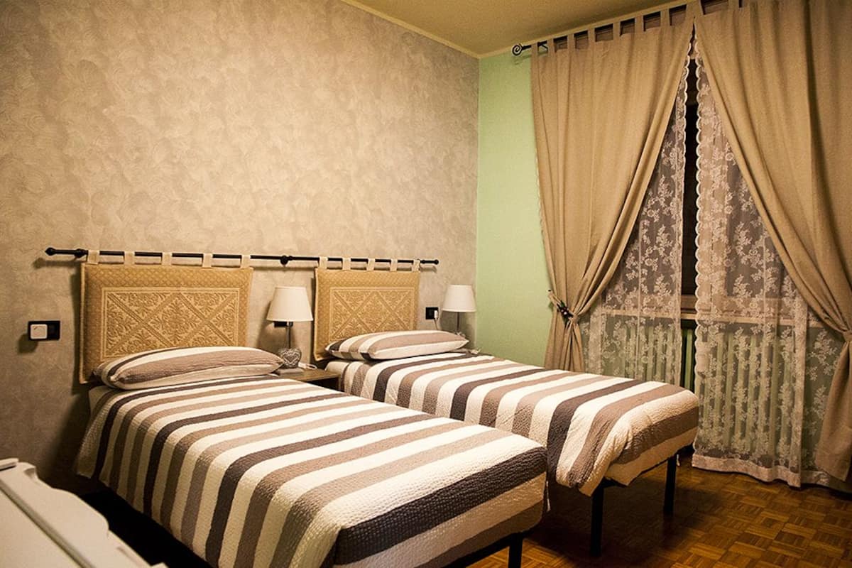 A hotel room with two beds, a nightstand, a lamp, and a window with curtains.