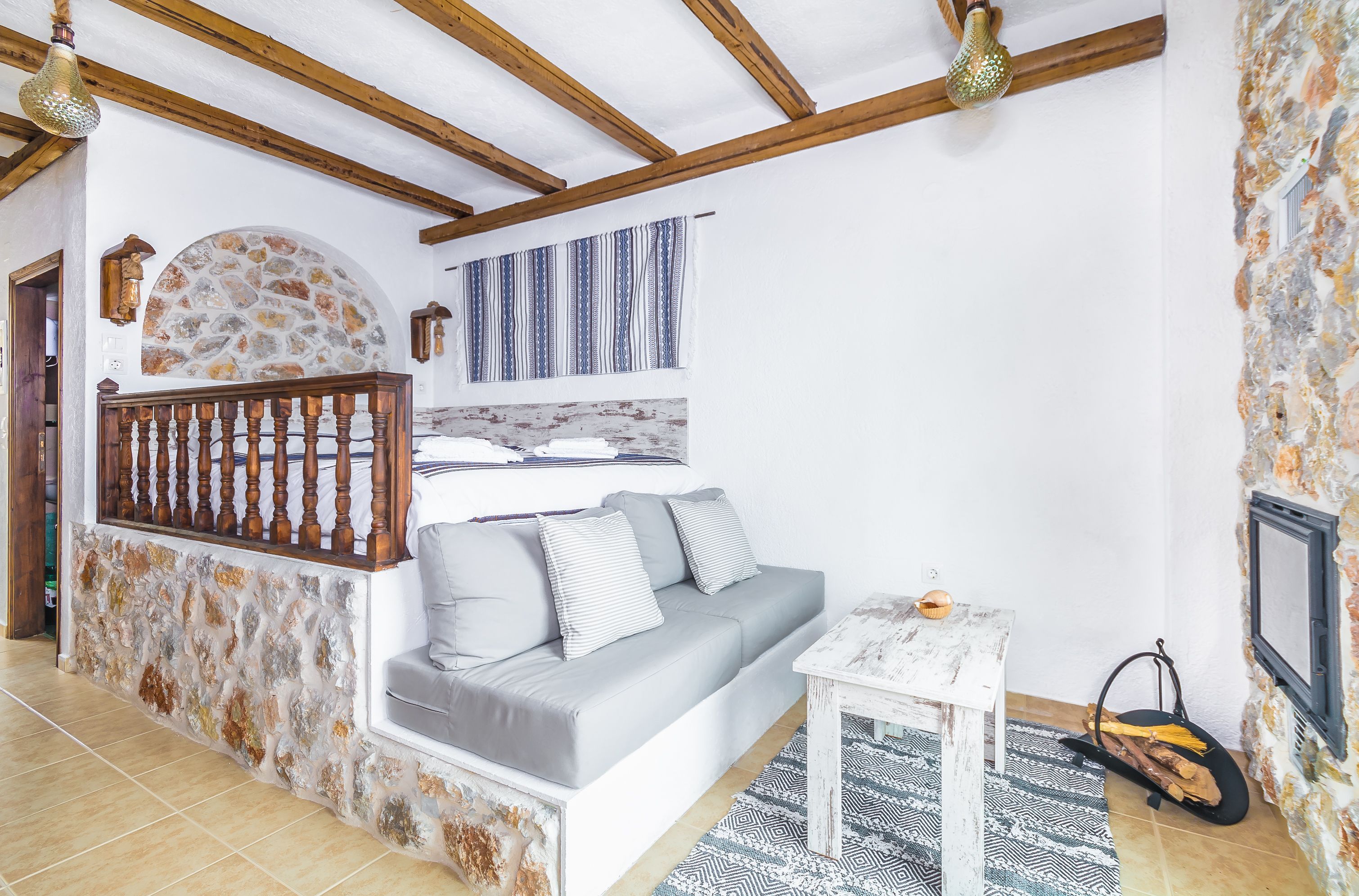 A cozy room with a stone wall, wooden ceiling beams, a grey sofa, a small white table, and a patterned rug.