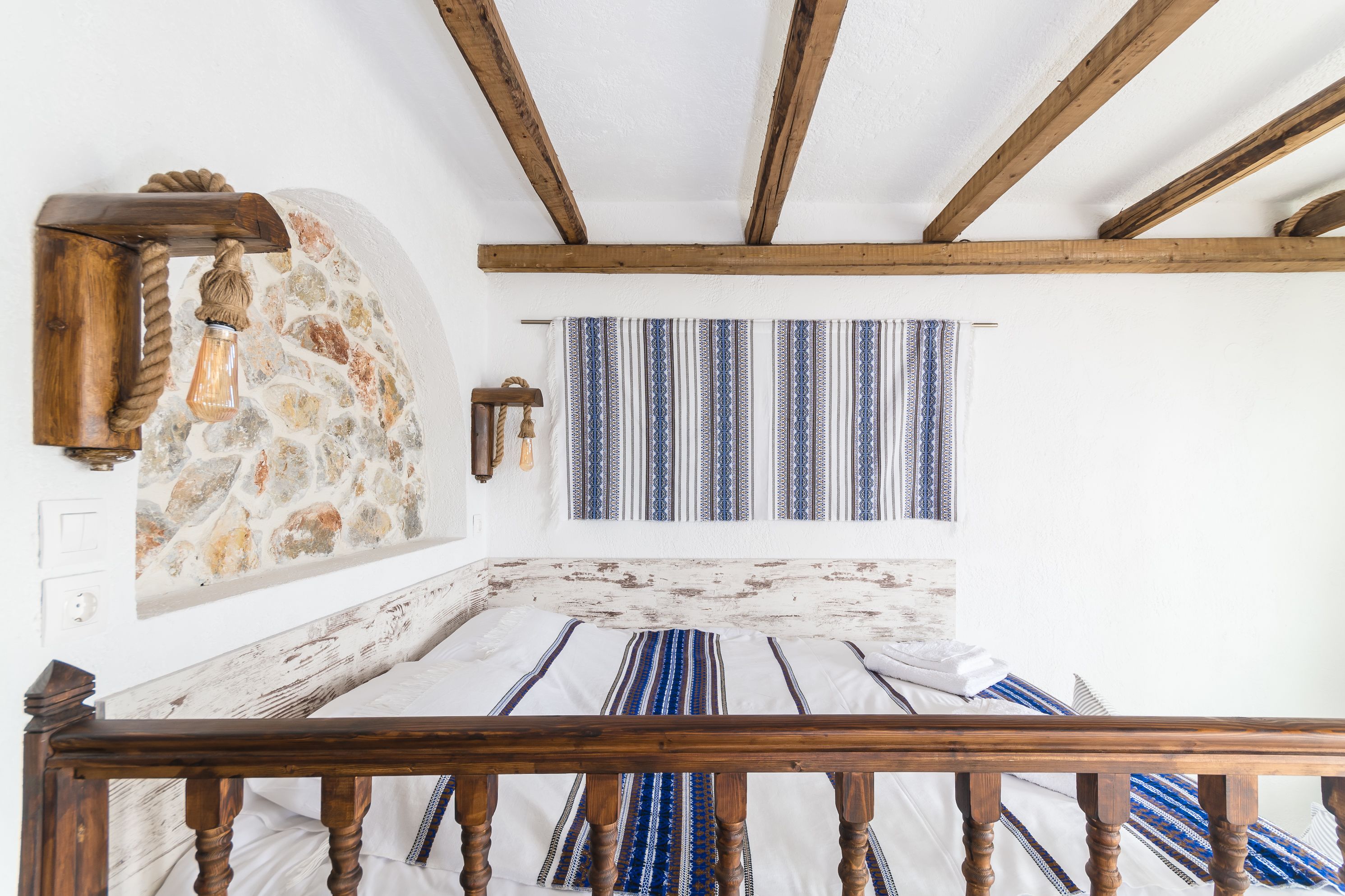A room with a stone wall, wooden beams, a bed with striped bedding, and a wooden railing.