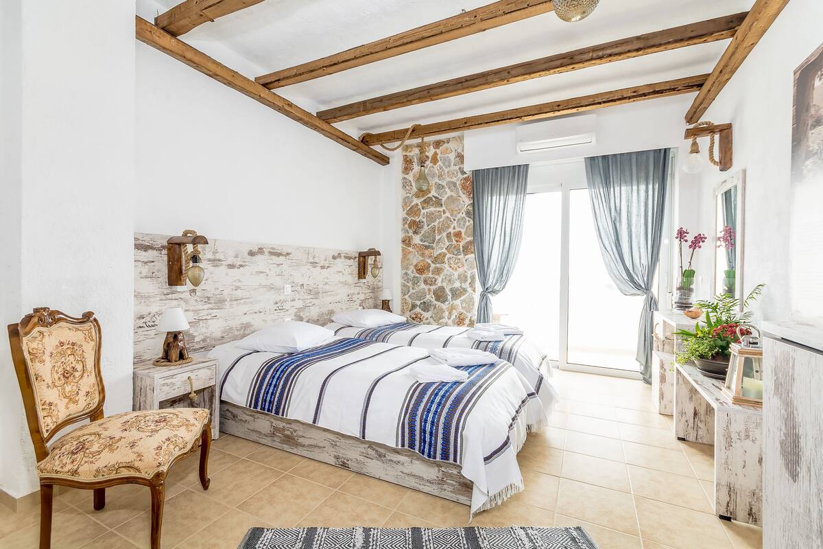 Ein Schlafzimmer mit Steinsandsteinwand, Holzbalken, einem Bett mit gestreifter Bettwäsche, einem Sessel, Nachttischen und einem Fenster mit Vorhängen.