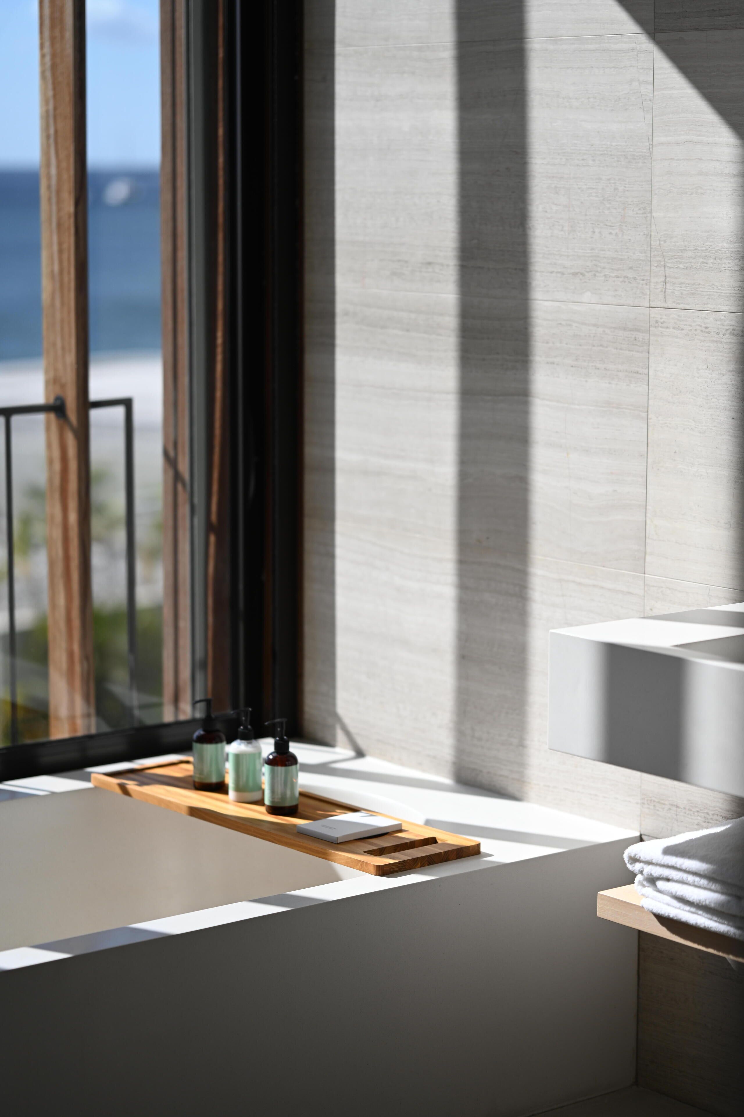 Un baño moderno con bañera, una bandeja de madera con botellas y vista al mar a través de un ventanal grande.