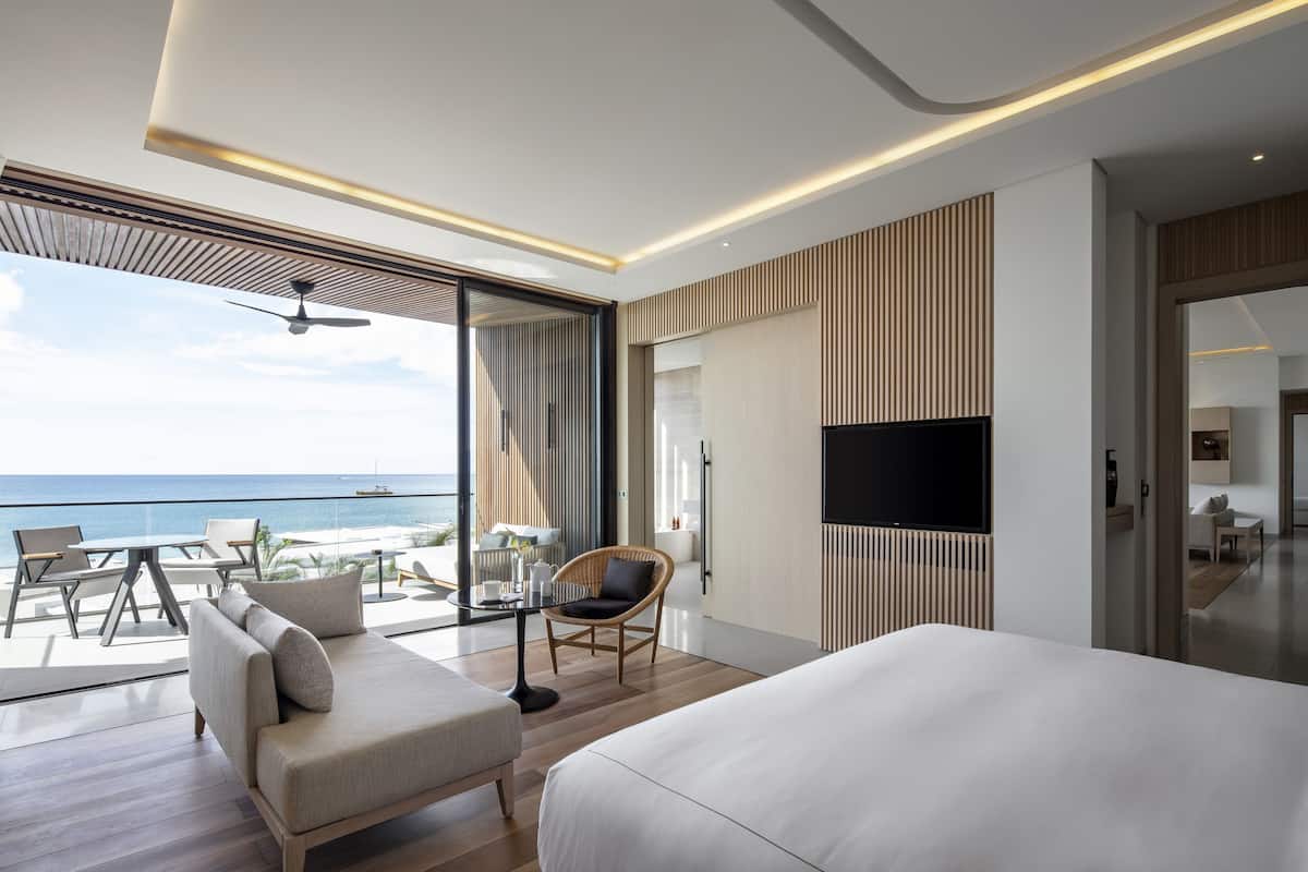 A modern hotel room with a large bed, a seating area with a table and chairs, a television, and a view of the ocean.