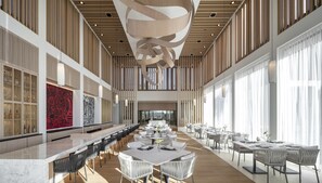 A spacious hotel lobby with a long reception desk, multiple dining areas with round tables, and a high ceiling with a unique light fixture.